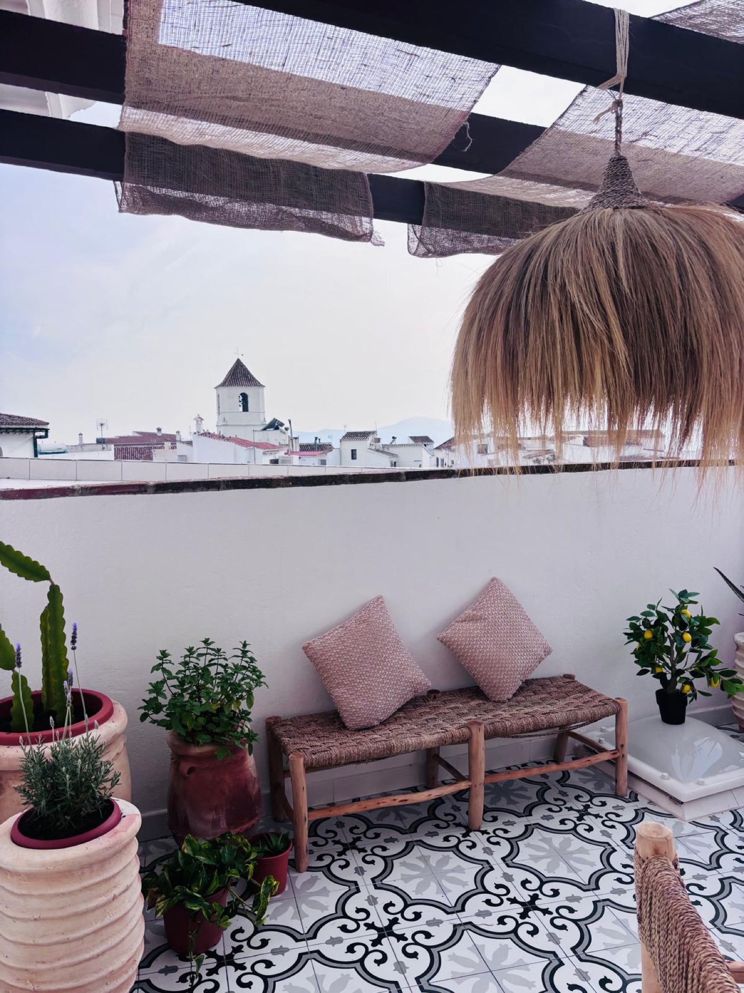 Rincón de terraza con banco de madera y enea con cojines rosas, macetas de barro con cactus y plantas, suelo de baldosa hidráulica y lámpara de fibra natural, con la torre de la iglesia del pueblo blanco al fondo.