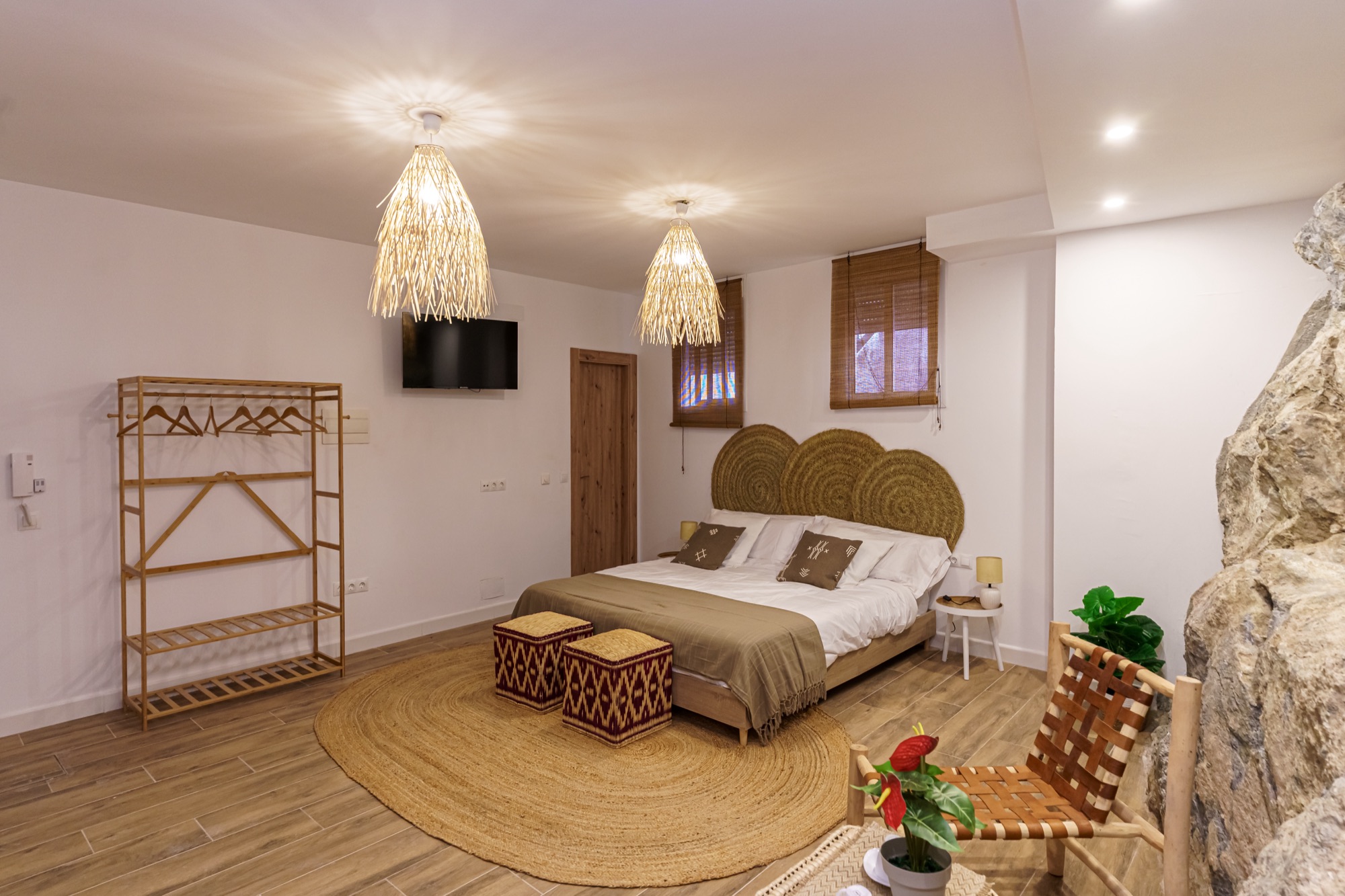 Panorámica del dormitorio de Maroma con las lámparas de fibra natural, televisor, burro de madera para la ropa, cama con cabecero de esparto y alfombra ovalada de yute.