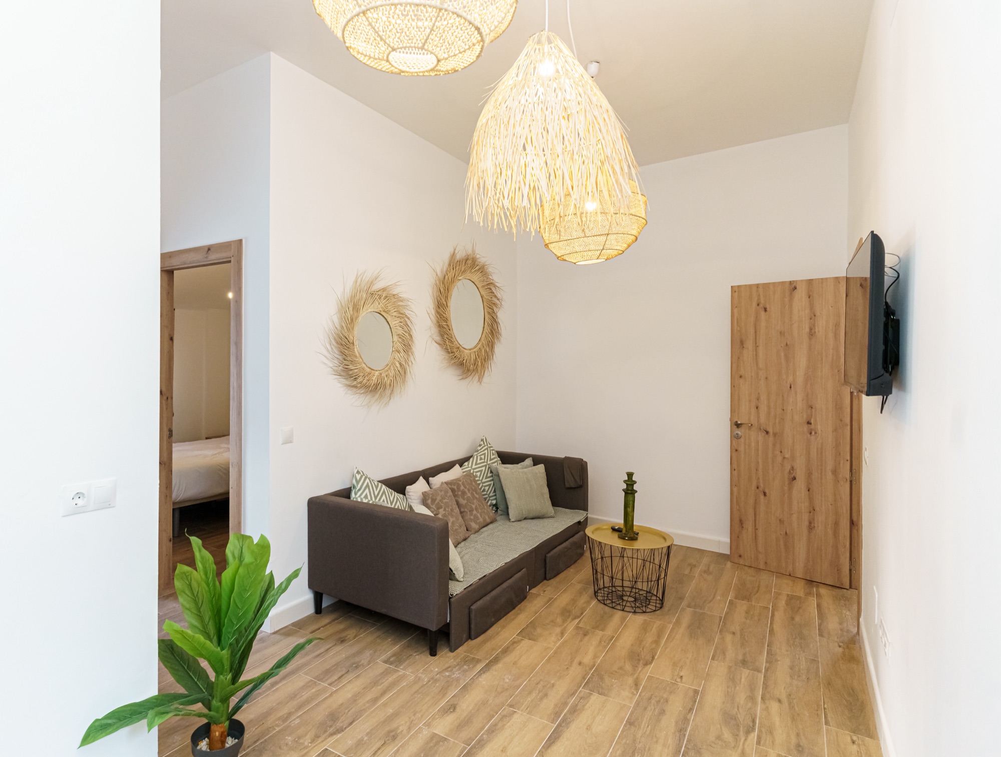 Salón del apartamento Olivo con sofá gris topo lleno de cojines en tonos salvia, lámparas colgantes de fibra natural, espejos de sol de rafia en la pared y puerta de madera clara.