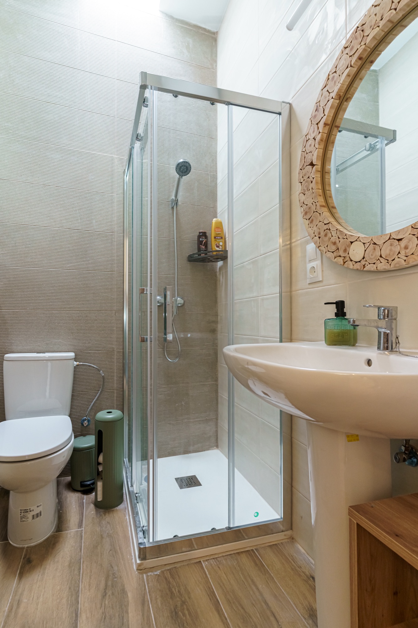Baño del apartamento Olivo con cabina de ducha acristalada, lavabo de pedestal, espejo redondo con marco de rodajas de madera y detalles en verde oliva.