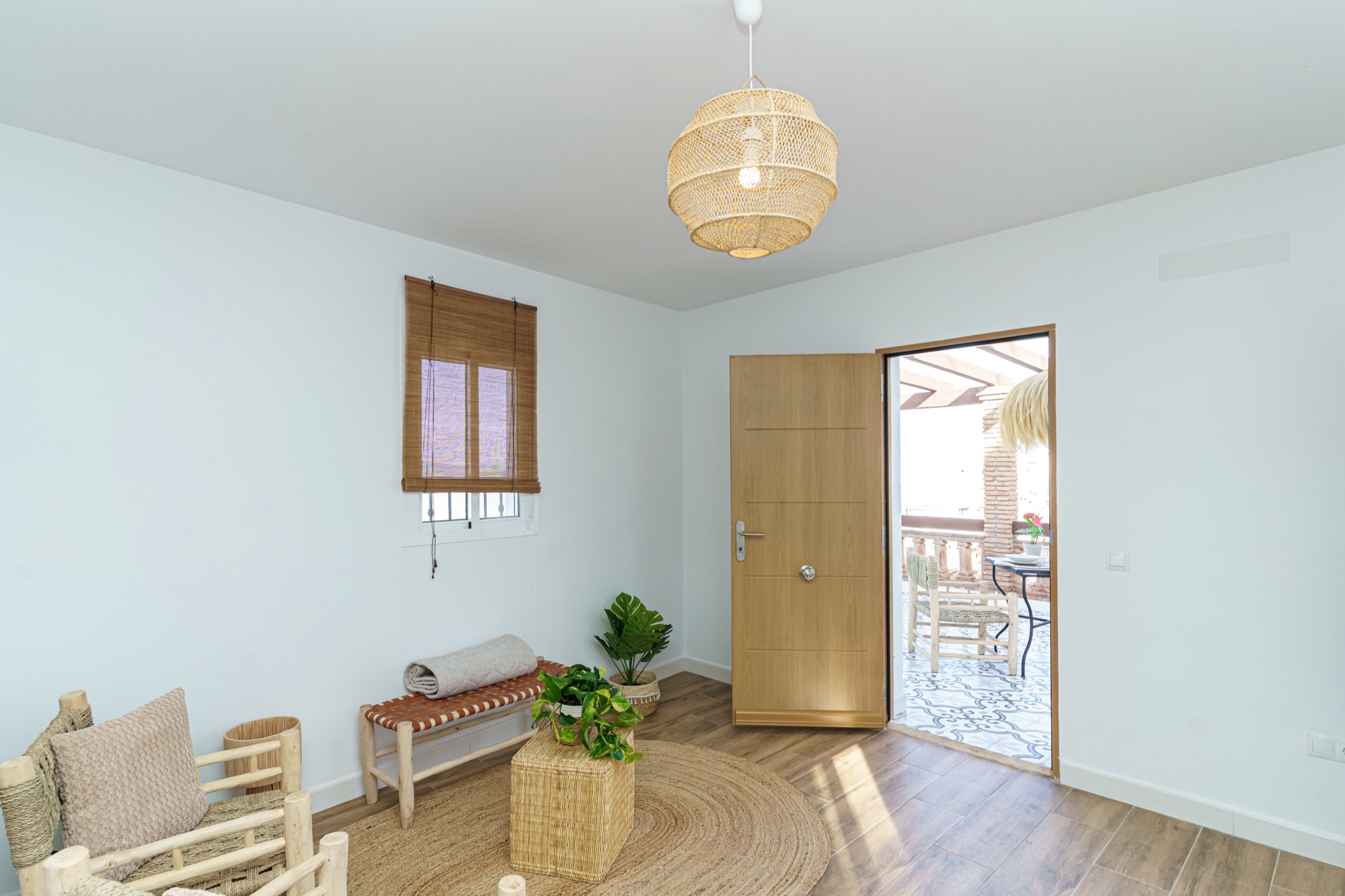 Salón con puerta de roble abierta hacia la terraza soleada, lámpara colgante de ratán, banco con esterilla enrollada y plantas sobre alfombra de yute.