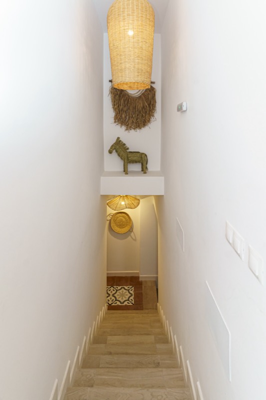 Escalera interior estrecha de paredes blancas vista desde arriba, decorada con una gran lámpara colgante de mimbre, un adorno de rafia, una figura de caballo tallado y sombreros de esparto.