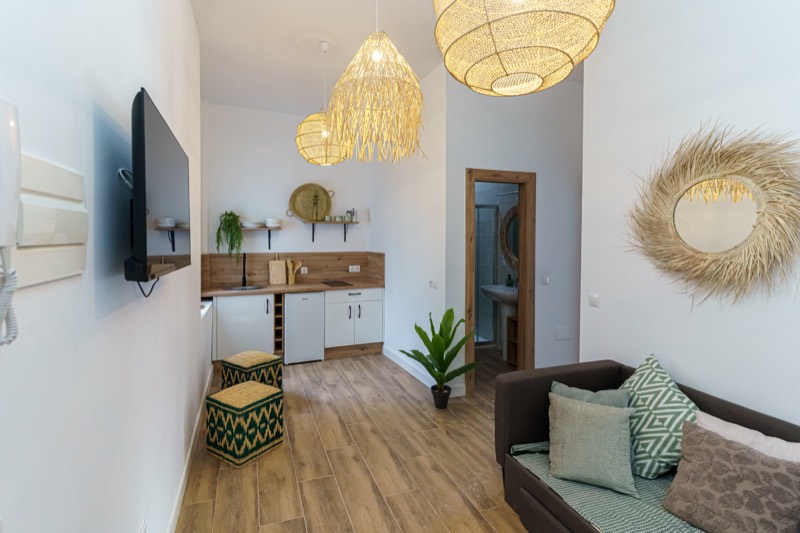 Vista general del salón con cocina abierta del apartamento Olivo: encimera de madera, muebles blancos, lámparas de fibra, sofá con cojines verdes y taburetes de mimbre estampados.