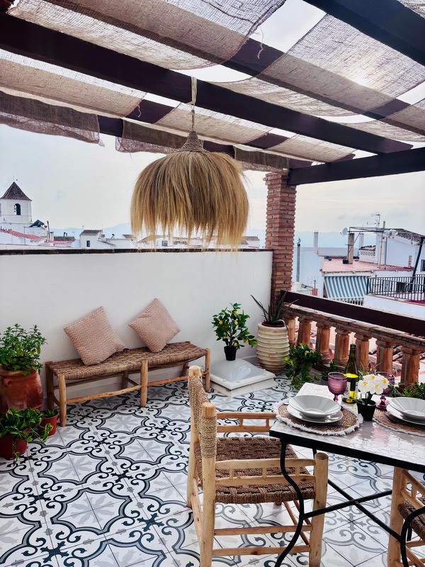 Terraza con pérgola de brezo y lámpara de rafia, suelo hidráulico blanco y negro, mesa de mosaico puesta y vistas a los tejados blancos y al campanario de Canillas de Aceituno.