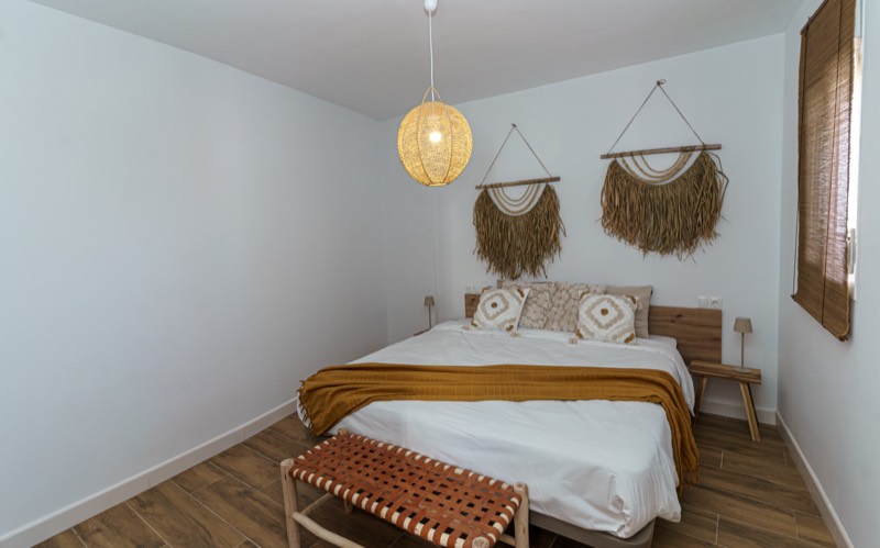 Dormitorio con cama doble vestida de blanco y manta mostaza, dos tapices de rafia sobre el cabecero de madera, lámpara de fibra natural y banqueta de tiras de cuero a los pies.